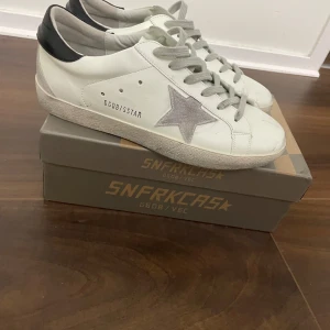 Vita sneakers från Golden Goose - Säljer ett par vita Golden Goose sneakers i bra skick. De har en grå stjärna på sidan och svarta detaljer vid hälen. Skorna är använda 2-3 gånger och väldigt fräscha skorna är även 100%äkta och har ett digitalt kvittobevis så passa på för det är även väldigt lågt pris för att vara Golden goose. Med skorna tillkommer även en box och en påse så passa på!!