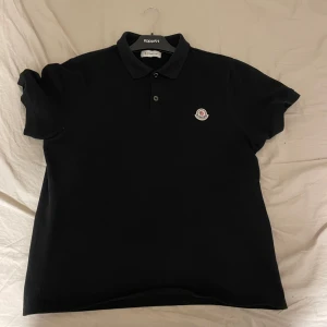 Svart pikétröja från Moncler - Säljer en stilren svart pikétröja från Moncler i storlek L. Tröjan har korta ärmar och en klassisk krage med knappar. Perfekt för både vardag och festliga tillfällen. Moncler-loggan är snyggt placerad på bröstet. Passar perfekt till jeans eller chinos!