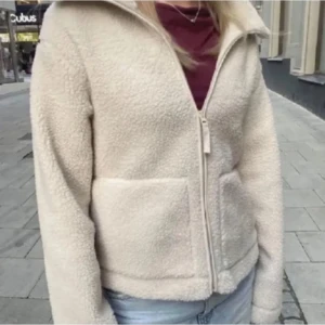 Beige teddyjacka med dragkedja - Mysig beige teddyjacka perfekt för kyliga dagar. Jackan har en dragkedja framtill och två praktiska fickor. Den är långärmad och har en hög krage för extra värme. Perfekt för höst och vinter!