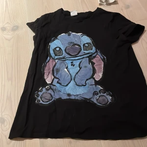 Svart t-shirt med Disney-motiv - Säljer en svart t-shirt med ett gulligt motiv av en blå karaktär från Disney. T-shirten är kortärmad och i bra skick. Perfekt för alla Disney-fans som vill ha något bekvämt och stilrent i garderoben!