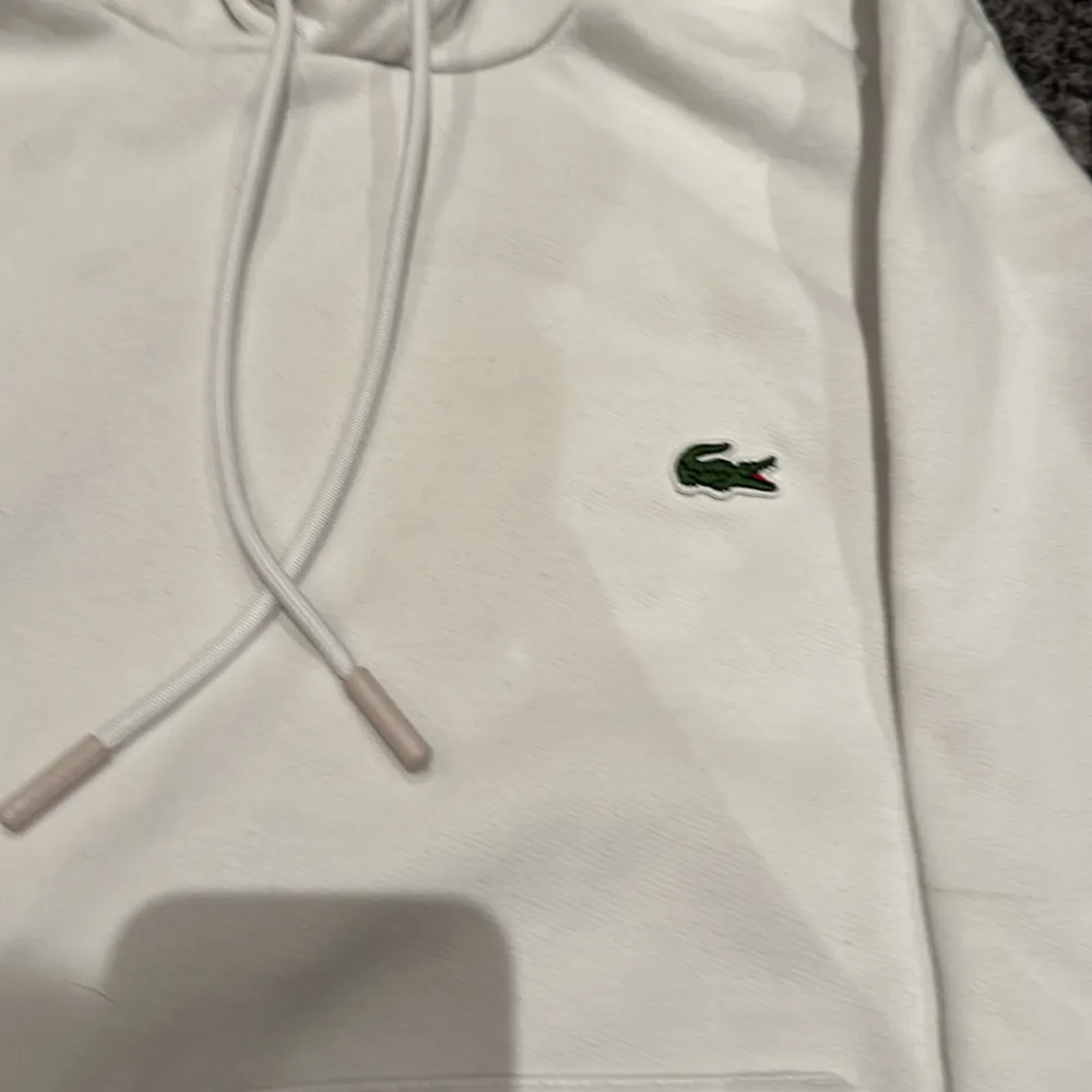 Säljer en stilren vit hoodie från Lacoste i klassisk passform. Den har en känguruficka och justerbar huva med dragsko. Perfekt för en avslappnad stil och passar till alla säsonger. Riktigt skön. 🐊. Tröjor & Koftor.