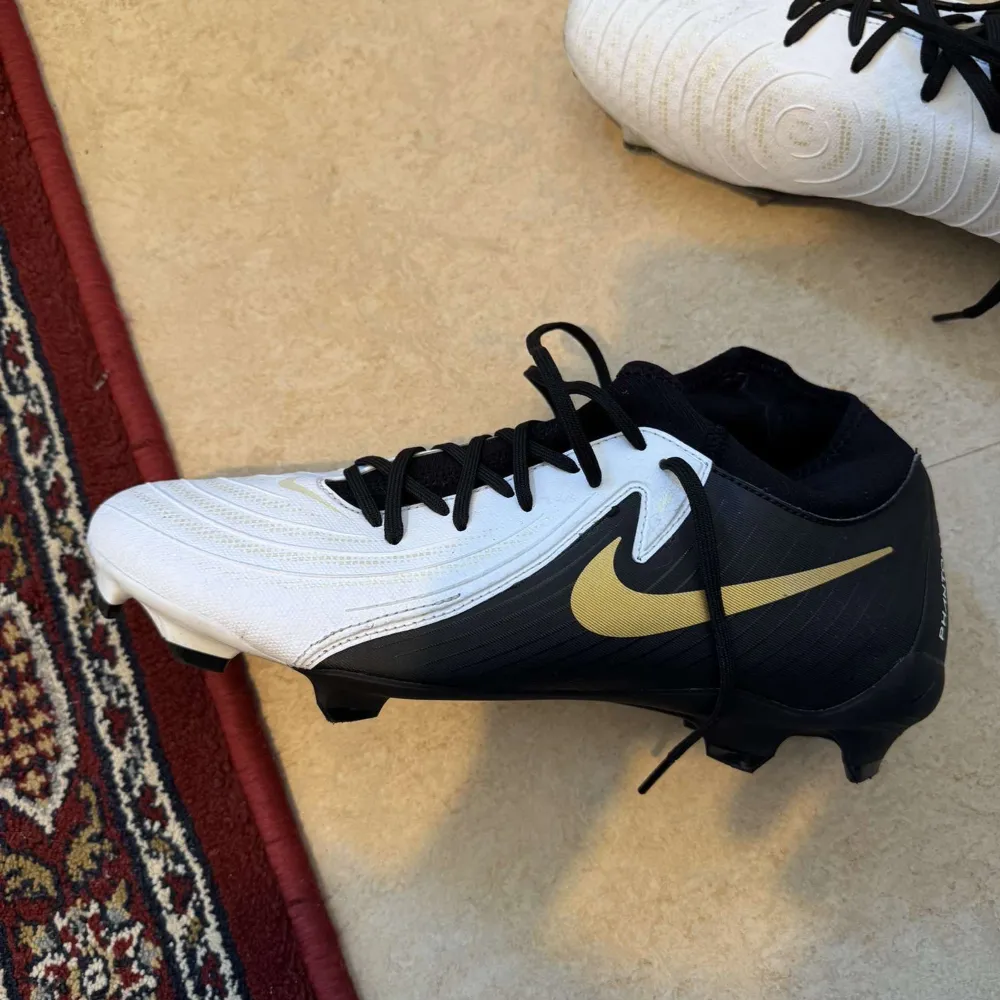 Säljer ett par Nike Phantom fotbollsskor i svart och vitt med gulddetaljer. Skorna har snörning och är i mycket bra skick. Perfekta för fotbollsspelare som vill ha stil och funktion på planen.. Skor.