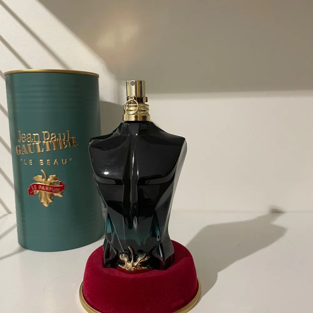 Säljer en cool parfymflaska formad som en manlig torso från Jean Paul Gaultier. Flaskan är mörkgrön och svart med en guldig spraytopp. Den kommer i en elegant grön metallförpackning med guldtext. Perfekt för den som vill ha en unik och stilren doftupplevelse. Flaskan står på en röd sammetsbas.. Perfume.