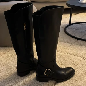 a Pair Svarta höga stövlar med dragkedja - Snygga svarta stövlar i skinn med dragkedja och spänne i guld. Perfekta för höst och vinter, dessa stövlar har en klassisk design som passar både till vardags och fest. De är i bra skick och redo att användas direkt. 