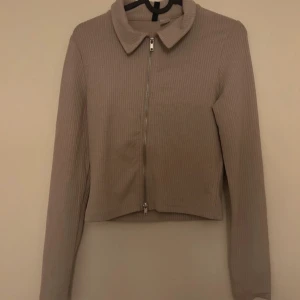 Beige ribbad tröja med dragkedja - Säljer en snygg beige ribbad tröja med dragkedja framtill. Den har en klassisk krage och långa ärmar, perfekt för både vardag och fest. Tröjan är croppad och ger en stilren look.