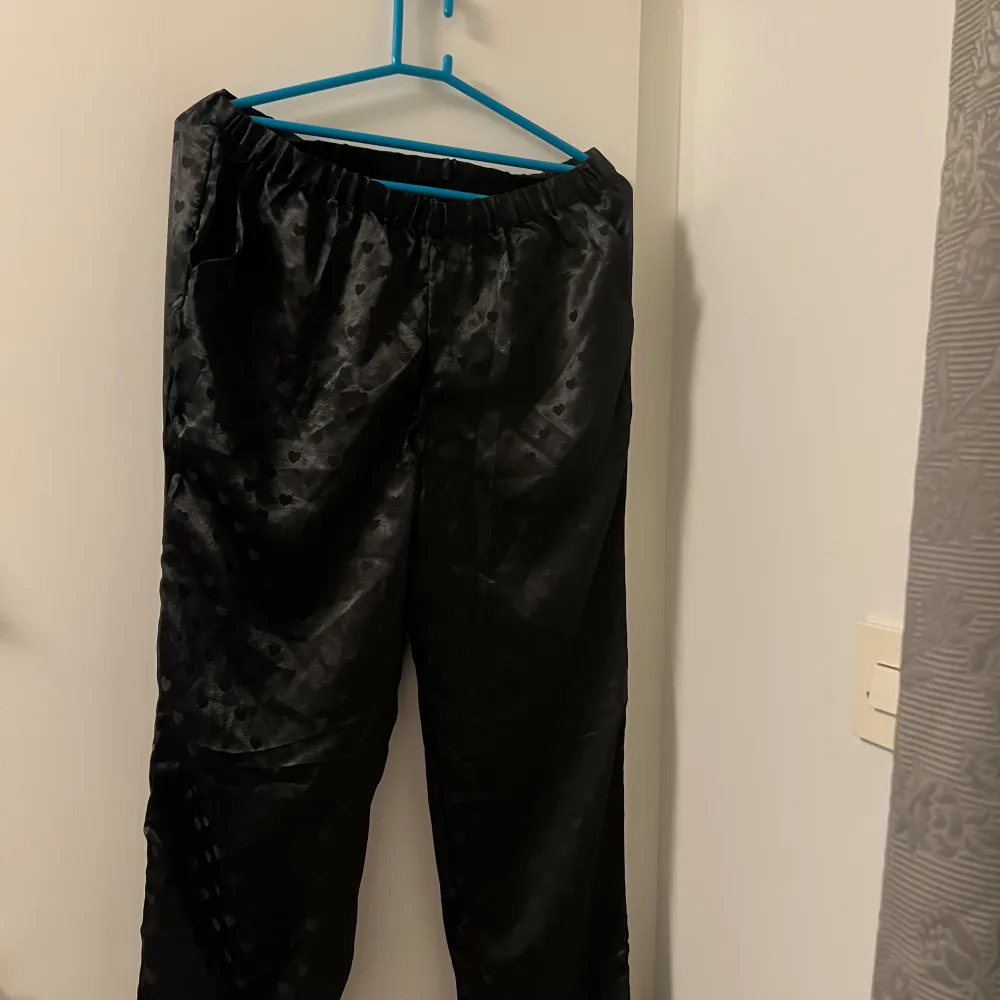 Säljer ett par svarta byxor i satin material med hjärtmönster från Boohoo 🎀 Byxorna har en loose passform och elastisk midja för extra komfort för era myskvällar. De är i mycket bra skick och redo för en ägare 🙏. Jeans & Byxor.