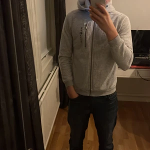 Grå hoodie från Sail Racing - Säljer en snygg grå hoodie från Sail Racing i bra skick. Den har en dragkedja framtill och en praktisk ficka med dragkedja på bröstet. Perfekt för både vardag och chill. Storleken är normal och den är supermjuk och bekväm att ha på sig.