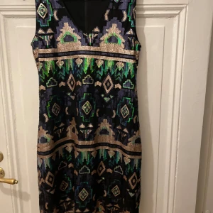 Glittrig ärmlös klänning - Säljer en superfin glittrig klänning med ett coolt geometriskt mönster i grönt, blått och silver. Den är ärmlös och har en v-ringad hals. Perfekt för festliga tillfällen när du vill sticka ut! Klänningen är i bra skick och redo för nya äventyr.
