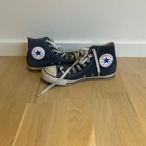 Blå Converse All Star - Säljer ett par klassiska blå Converse All Star i bra skick. De har en hög modell med snörning och den ikoniska loggan på sidan. Strlk 40. Det är använda och har lite märken men de går säkert bort när man tvättar de!