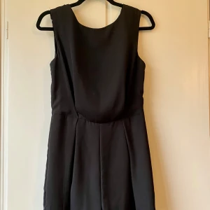 Svart ärmlös byxdress - Säljer en elegant svart ärmlös byxdress i mycket bra skick. Den har en stilren design med en djup v-ringad rygg och dragkedja. Perfekt för festliga tillfällen eller en utekväll. 