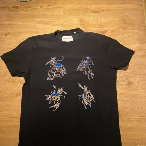 Svart t-shirt från Our Legacy - Säljer en svart t-shirt från Our Legacy i storlek 52. Den har ett coolt broderat mönster med cowboys i olika färger på framsidan. Perfekt för en avslappnad stil med en touch av western. T-shirten är kortärmad och gjord i ett mjukt material.