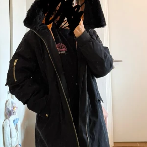 Svart parkajacka med fuskpäls från Whyred - Säljer en svart parkajacka från Whyred med fuskpäls på huvan. Jackan har en dragkedja i guld och praktiska fickor. Perfekt för kalla dagar med sin långa passform och varma foder. Den är i mycket bra skick och redo för vinterns äventyr!