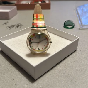 Färgglad klocka med rutigt mönster Swatch - Säljer en supercool klocka med ett färgglatt rutigt mönster i rött, gult, grönt och orange. Klockan har en vit urtavla med färgglada siffror och ett transparent armband. Perfekt för att liva upp vilken outfit som helst!