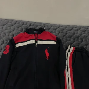 Polo Ralph Lauren Dress - Säljer en snygg svart hoodie från Polo Ralph Lauren med röda och vita detaljer. Den har en dragkedja framtill och ett stort rött logotryck på bröstet. Perfekt för en sportig look eller chill dagar. Storlek är inte angiven, men den ser ut att vara i bra skick. Pris kan diskuteras.