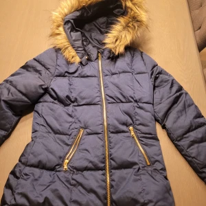 Mörkblå dunjacka från H&M - Säljer en snygg mörkblå dunjacka från H&M med fuskpäls på huvan. Jackan har dragkedja och två praktiska fickor med guldfärgade dragkedjor. Perfekt för kalla höstdagar.