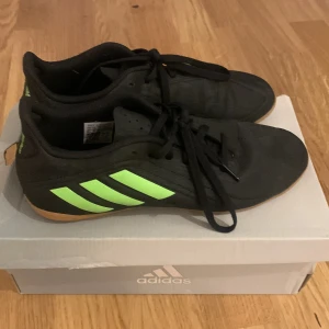 Svarta Adidas fotbollsskor med gröna detaljer - Säljer ett par svarta Adidas fotbollsskor i mycket bra skick. De har gröna detaljer och klassiska tre ränder på sidan. Skorna är perfekta för inomhusfotboll och har en bekväm passform med snörning. Använda endast ett fåtal gånger.