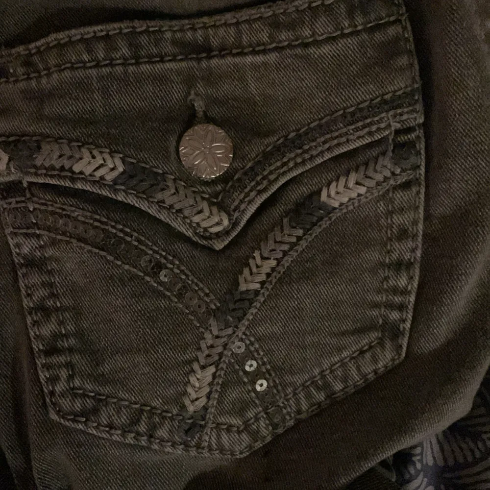 Säljer ett par snygga grå jeans med unika detaljer på bakfickan. De har en cool knapp och flätade sömmar som ger en extra touch. Perfekt för vardagsbruk eller en utekväll. De är bekväma och stilrena, ett måste i garderoben!. Jeans & Byxor.