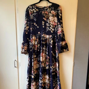 Blommig långklänning - Säljer en superfin blommig långklänning i mörkblått med vackra blommor i rosa och beige toner. Klänningen har långa ärmar och en luftig passform. Materialet känns mjukt och bekvämt, och den har en romantisk stil som passar till både vardag och fest.