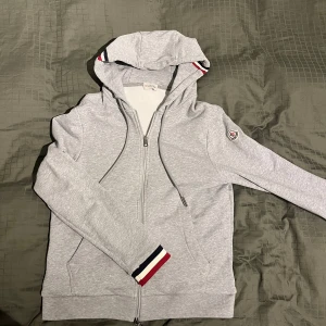 Moncler zip hoodie grå - Säljer en snygg grå hoodie från Moncler i mycket bra skick. Den har en dragkedja framtill och coola detaljer med röd och svart rand vid ärmslut och huva. Perfekt för höst och vår när du vill vara både bekväm och stilren. Priset kan diskuteras vid snabb affär!