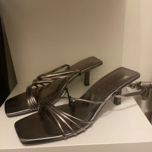 Klackar - Snygga silvriga/champagnefärgade sandaler med klack och smala remmar som korsar över foten. Perfekta för en utekväll eller fest. De har en modern, fyrkantig tå och en elegant design som verkligen sticker ut. Passar bra till både klänning och byxor. Aldrig använda