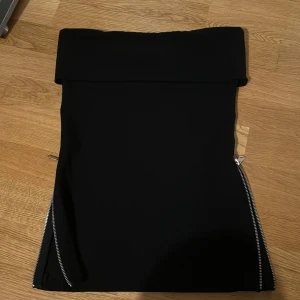 Svart tubtopp med dragkedjor från Zara - Snygg svart tubtopp från Zara i storlek XS. Toppen har en tight passform och är försedd med coola dragkedjor på sidorna för en edgy look. Dragkedjorna funkar perfekt!  Dubbeltyg vid bysten, fin detalj och bra stöd. Perfekt för både fest och vardag. 