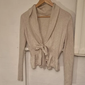 Beige stickad kofta med knyt - beige stickad kofta med knyt framtill. Långa ärmar och en volangdetalj vid nederkanten. Storlek S och är i Bra skick 