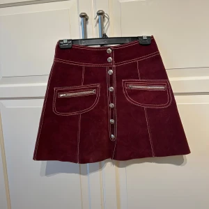 Vinröd mockakjol med knappar - Säljer en superfin vinröd äkta mockakjol med snygga silverknappar framtill. Kjolen har två praktiska fickor med dragkedjor och kontrastsömmar som ger en cool look. Perfekt för höst och vinter, och funkar lika bra till vardags som till fest. 👜✨