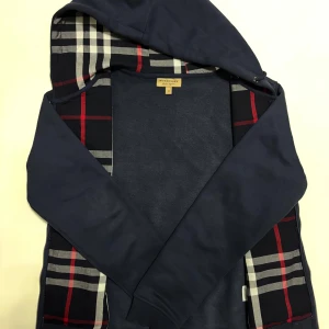 Marinblå hoodie från Burberry - Säljer en rare marinblå hoodie från Burberry med klassiskt rutigt mönster i huvan. Tröjan har dragkedja och fickor framtill. Har använt den kanske 6-7 ggr bara. Just den färgen marinblå är svår att få tag i så passa på. Storleken är M men kan även passa någon med S. Skickas inom 24 timmar.