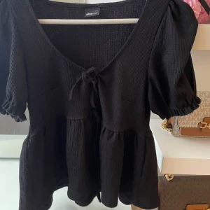 Svart blus från Gina Tricot - Snygg svart blus från Gina Tricot med puffiga kortärmar och knytning framtill. Den har en v-ringad design och är tillverkad i ett ribbat material som ger en fin struktur.