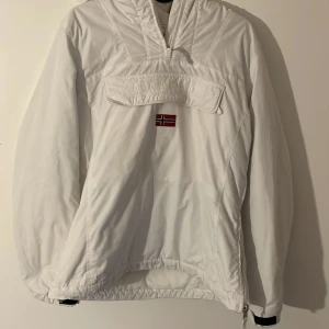 Vit jacka från Napapijri - Säljer en stilren vit jacka från Napapijri med norsk flagga på bröstet. Jackan har en praktisk huva och dragkedja framtill. Perfekt för kyligare dagar.