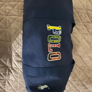 Blå axelväska från Polo Ralph Lauren - Snygg blå axelväska från Polo Ralph Lauren med färgglada bokstäver på framsidan. Väskan har en broderad logotyp i vitt och justerbar axelrem. Perfekt för en stilren look.