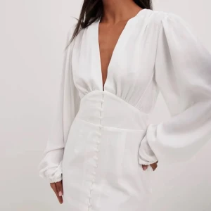 Vit klänning med djup v-ringning - Elegant vit klänning med djup v-ringning och långa ärmar. Klänningen har knappar framtill och en figursydd passform som ger en stilren look. Helt ny oanvänd 299