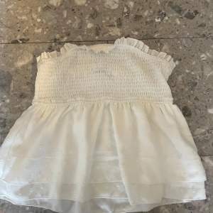 Säljer en söt vit axelbandslös topp med smockdetaljer och volangkant. Toppen har ett skirt lager med små prickar som ger en fin textur. Perfekt för varma sommardagar!