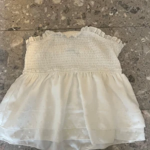 Vit axelbandslös topp - Säljer en söt vit axelbandslös topp med smockdetaljer och volangkant. Toppen har ett skirt lager med små prickar som ger en fin textur. Perfekt för varma sommardagar!