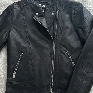 Svart skinnjacka från KappAhl - Snygg svart skinnjacka från KappAhl med dragkedja och stilren design. Jackan har en asymmetrisk dragkedja framtill och två fickor med dragkedjor. Storlek 170 men skulle säga att det är en XS. Den är helt oanvänd.
