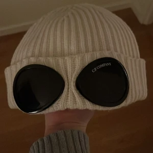 Vit ribbad mössa med glasögondetalj från C.P. Company - Snygg vit ribbad mössa från C.P. Company med unika glasögondetaljer framtill. Perfekt för att ge din outfit en cool touch.
