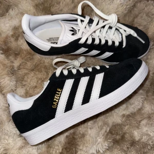 Svarta Adidas Gazelle sneakers - Säljer ett par klassiska svarta Adidas Gazelle sneakers med vita detaljer och snörning. Skorna har den ikoniska tre-randiga designen och är tillverkade i mocka med en vit sula. Skorna är använda en gång, men ser fortfarande helt nya ut. Svarta snören medföljer vid köp.