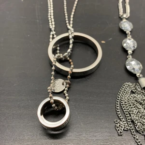 Två halsband med ring- och tofsdetaljer - Säljer två stilrena halsband. Det ena har en ring med ingraverad text och kulkedja, 42 cm långt. Det andra har glittrande stenar och en tofs av kedjor, 50 cm långt. Perfekta för att ge en elegant touch till din outfit.