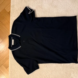 Svart pikétröja med vita detaljer - Snygg svart pikétröja med vita detaljer på kragen. Perfekt för en stilren look. Kortärmad och tillverkad i ett bekvämt material. Storlek M. Mycket bra skick.