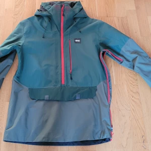 Grön skidjacka / snowboardjacka från Picture - Snygg grön skidjacka snowboardjacka från franska märket Picture. Med röda dragkedjor och justerbar huva. Jackan har en praktisk design med flera fickor, bland annat för liftkortet i ärmen. Anorak Om öppna via en dragkedja i sidan. 