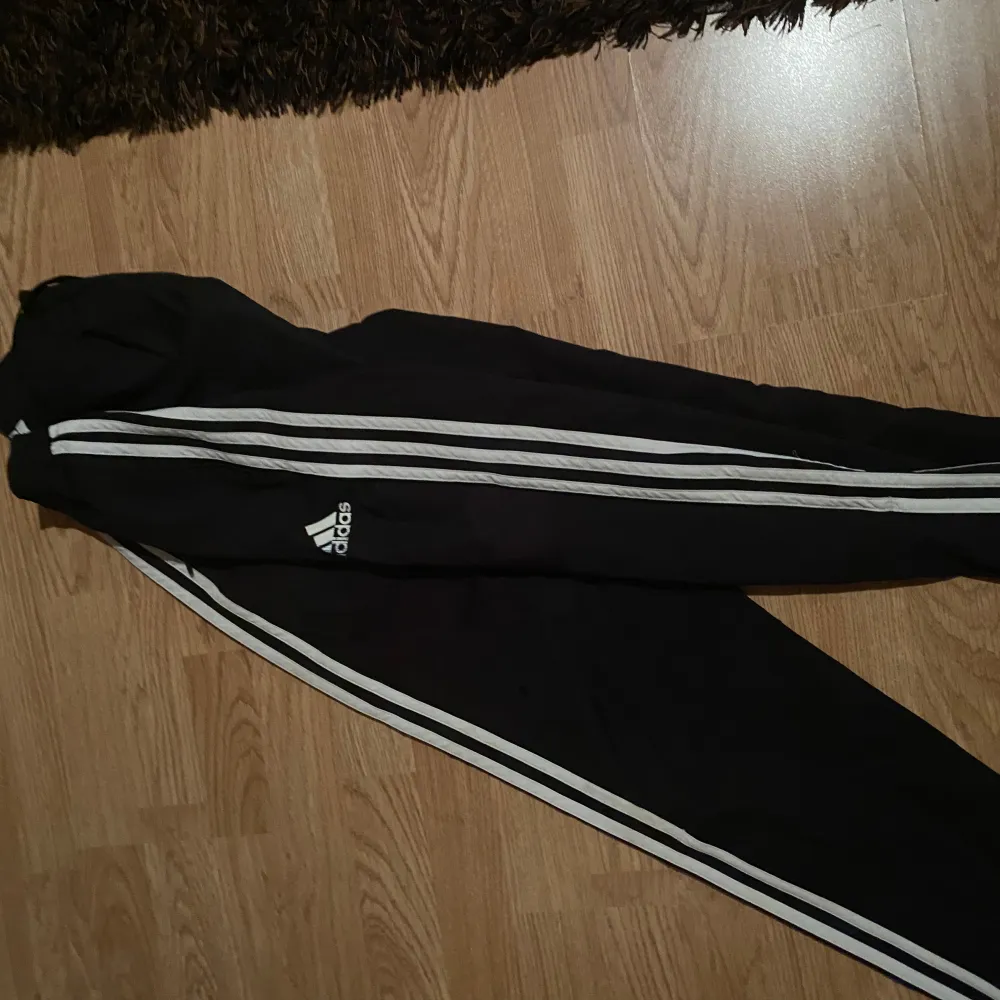 Säljer ett par svarta träningsbyxor från Adidas med vita ränder längs sidorna. Byxorna har en elastisk midja med snörning och är perfekta för sport och träning.. Farkut & Housut.