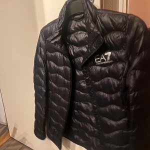 Svart dunjacka från Emporio Armani - Snygg svart dunjacka från Emporio Armani EA7 med quiltad design. Jackan har en dragkedja framtill och är perfekt för kyligare väder. Den har en stilren look med långärmade ärmar och en hög krage.