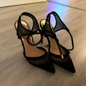 Svarta pumps med spetsig tå - Eleganta svarta pumps med spetsig tå och dekorativa detaljer. Skorna har en stilren design med en smal klack och knytning runt vristen.