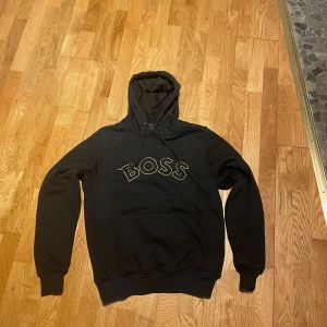 Svart hoodie från BOSS - Snygg svart hoodie från BOSS med logga framtill. Den har en justerbar huva med snören och en stor ficka på magen. Perfekt för en avslappnad stil.