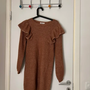 Brun stickad klänning från H&M - Säljer en mysig brun stickad klänning från H&M med volangdetaljer vid axlarna. Klänningen har långa ärmar och en rund halsringning, perfekt för kyligare dagar.