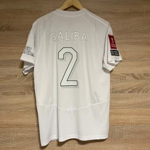 FA Cup Arsenal Saliba L - Säljer en vit Arsenal fotbollströja från Adidas med spelarnamnet Saliba och nummer 2 på ryggen. Tröjan har korta ärmar och tryck på ärmarna med Emirates FA Cup och No room for racism. Perfekt för fans av Arsenal och Saliba! Storlek L