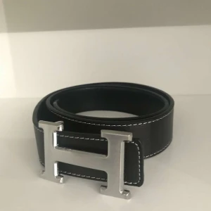 Svart skärp med silverfärgat H-spänne - Snyggt svart skärp med ett stort silverfärgat H-spänne. Skärpet har vita sömmar som ger en stilren kontrast. Perfekt för att lyfta vilken outfit som helst.