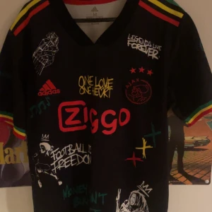 Svart fotbollströja från Adidas - Säljer en svart fotbollströja från Adidas med färgglada detaljer och tryck. Tröjan har korta ärmar och texten 'One Love One Heart' samt 'Live the Life You Love' tryckt på framsidan och baksidan. Perfekt för fotbollsfans som gillar unika och färgglada plagg.