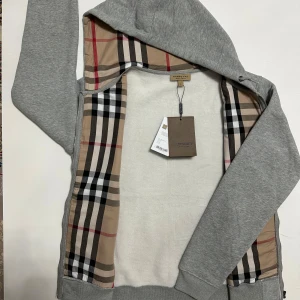  Burberry Kofta  - Säljer en stilren grå huvtröja från Burberry med klassiskt rutigt foder. Tröjan har dragkedja och en bekväm huva. Perfekt för en avslappnad och trendig look. Storleken är L. Skickas inom 24 timmar.