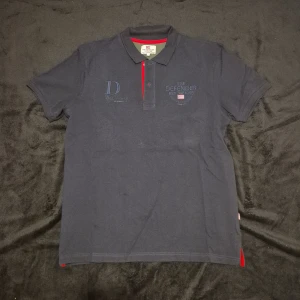 Mörkblå pikétröja med röda detaljer - Snygg mörkblå pikétröja från The Defender med röda detaljer vid knapparna och kragen. Tröjan har korta ärmar och en klassisk pikékrage. Perfekt för en stilren och avslappnad look.